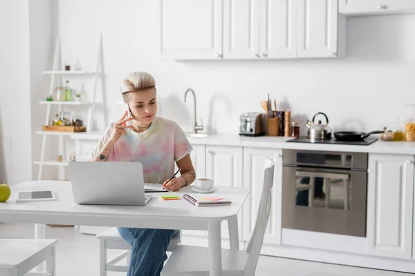 Young woman talking on smartphone and writing in notebook near laptop in kitchen — Stockfoto