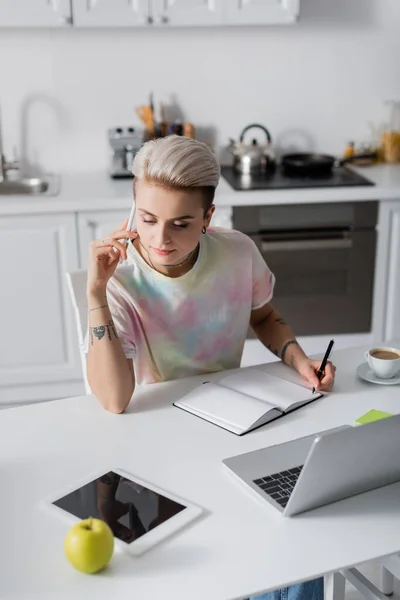 Donna elegante che scrive in notebook vuoto e parla sul cellulare vicino a laptop e tablet digitale — Foto stock