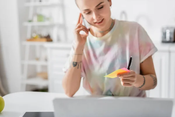 Mujer positiva sosteniendo notas adhesivas mientras habla en el teléfono inteligente cerca de la computadora portátil - foto de stock