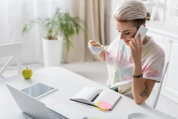 Trendy woman with coffee cup talking on smartphone near empty notebook, laptop and digital tablet — Photo de stock