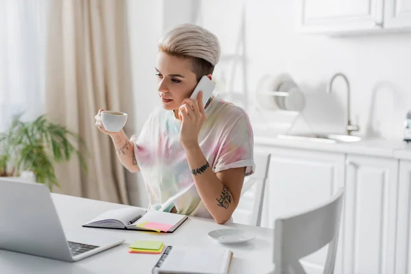 Tattooed woman holding coffee cup during conversation on smartphone near laptop and notebooks — Stock Photo