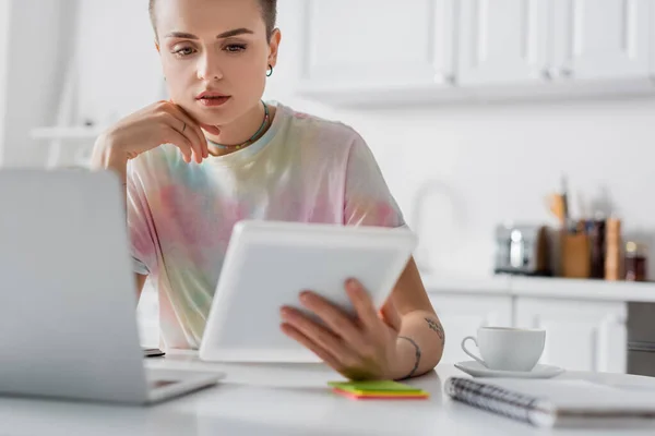 Усвідомлена жінка дивиться на цифровий планшет біля розмитого ноутбука на кухні — стокове фото