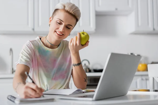 Mulher sorridente com maçã fresca escrita em notebook borrado perto de laptop — Fotografia de Stock