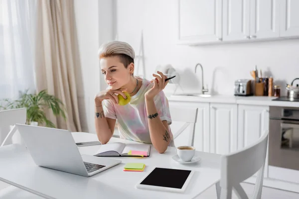 Татуированная женщина с яблоком и ручкой возле ноутбука, цифровой планшет и кофейная чашка — стоковое фото