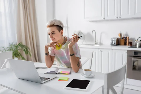 Mujer tatuada con manzana y pluma cerca de la computadora portátil, tableta digital y taza de café - foto de stock