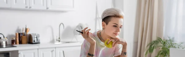 Tattooed woman with trendy hairstyle holding pen and fresh apple in blurred kitchen, banner — Foto stock