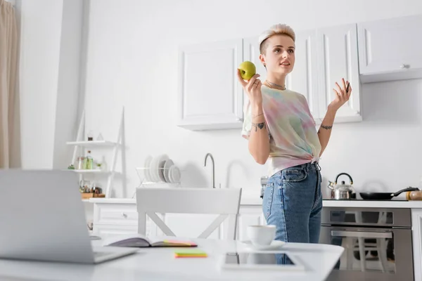 Mulher sorrindo de pé com maçã e caneta perto de laptop borrado e tablet digital na mesa da cozinha — Fotografia de Stock