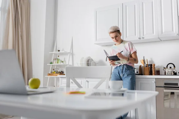 Jeune femme écrivant dans un carnet près d'un ordinateur portable sur une table de cuisine au premier plan flou — Photo de stock