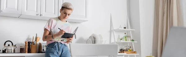 Jovem com penteado na moda escrevendo em notebook na cozinha em primeiro plano borrado, banner — Fotografia de Stock