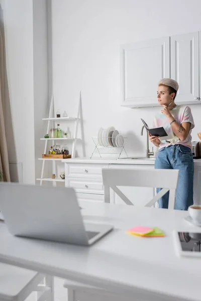 Pensive woman with notebook looking away in kitchen near blurred laptop on table — Photo de stock