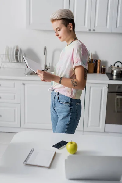 Mulher tatuada de pé com notebook vazio perto de maçã, smartphone e laptop na mesa da cozinha — Fotografia de Stock