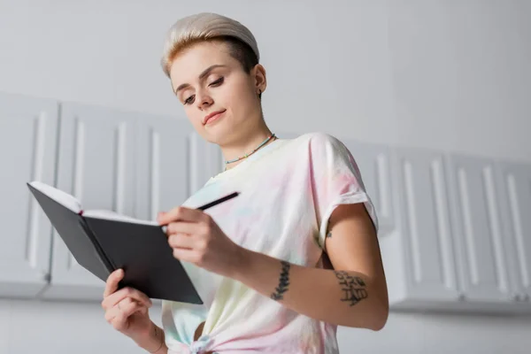 Low angle view of woman with trendy hairstyle writing in notebook in kitchen — Stock Photo