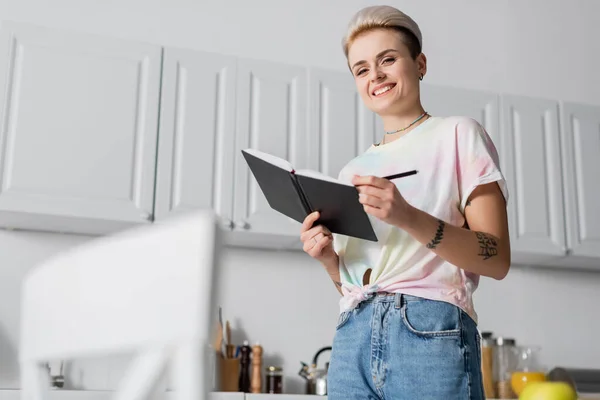 Low angle view of happy woman with notebook and pen looking at camera in kitchen — Stockfoto