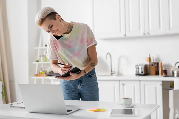 Donna sorridente con notebook che parla su smartphone vicino a laptop e tazza di caffè — Foto stock