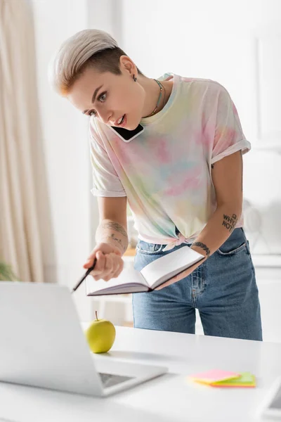 Junge Frau spricht mit Notizbuch und Stift auf Handy neben verschwommenem Laptop — Stockfoto