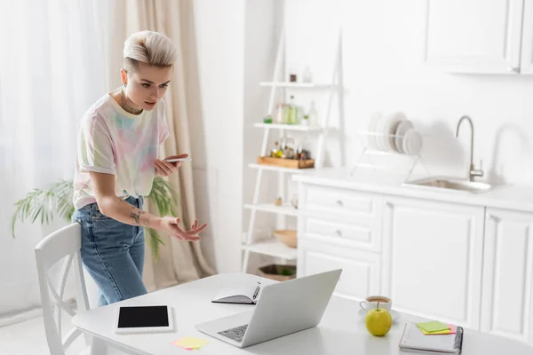 Trendy woman with mobile phone pointing with hand during video call on laptop in kitchen — Fotografia de Stock