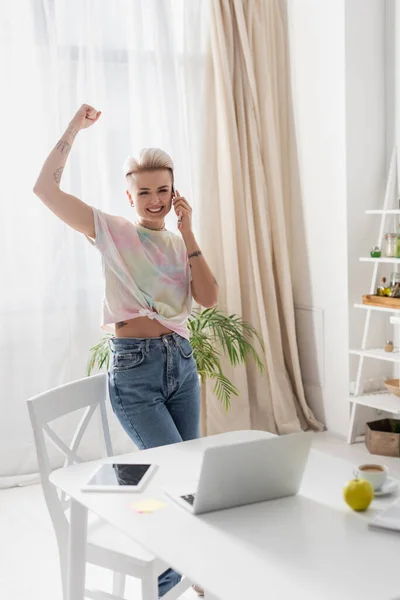 Aufgeregte Frau zeigt Triumphgeste, während sie in der Küche neben dem Laptop mit dem Smartphone spricht — Stockfoto