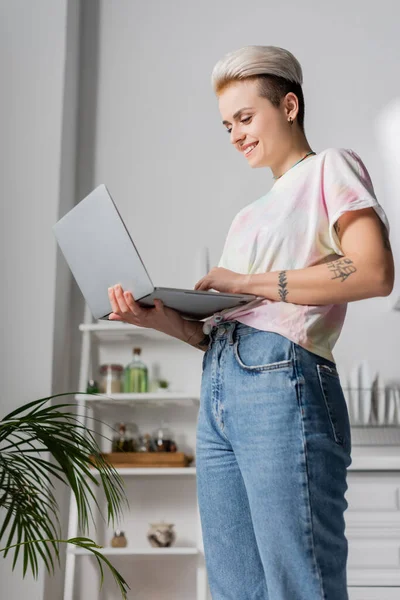 Щаслива татуйована жінка з модною зачіскою, що стоїть з ноутбуком вдома — стокове фото