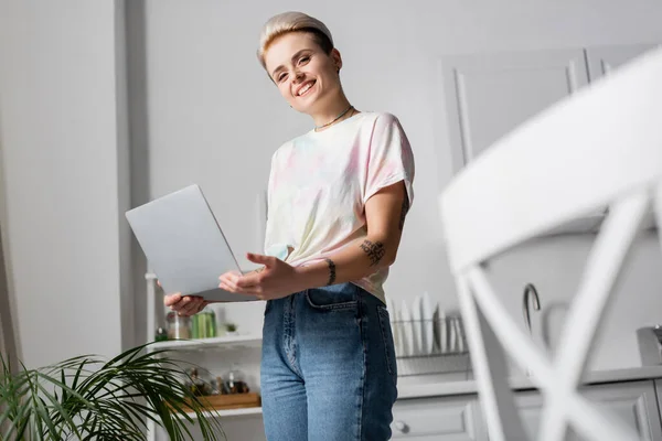 Femme tatouée gaie regardant la caméra tout en se tenant debout avec ordinateur portable à la maison — Photo de stock