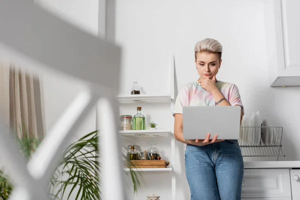 Thoughtful woman thinking near laptop in kitchen on blurred foreground — стоковое фото