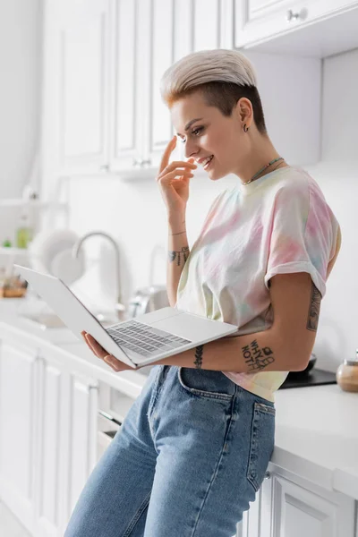 Femme gaie geste tout en se tenant debout avec ordinateur portable dans la cuisine — Photo de stock