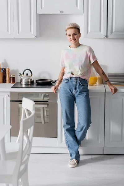 Visão de comprimento total da mulher sorridente em jeans olhando para a câmera na cozinha — Fotografia de Stock