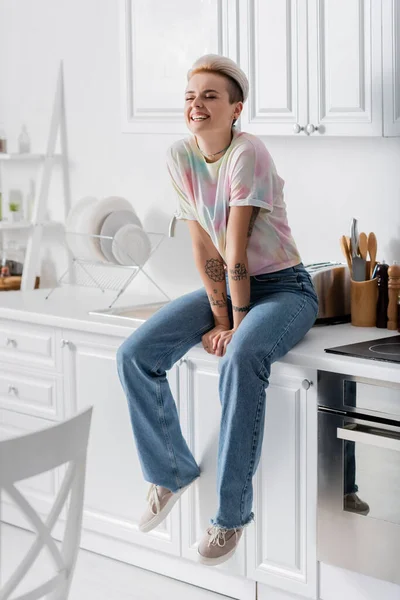 Mujer alegre y de moda con los ojos cerrados sentado en la encimera de la cocina - foto de stock