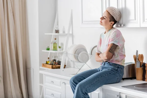 Verträumte und lächelnde Frau, die mit verschränkten Armen auf einer Arbeitsplatte in der Küche sitzt und wegschaut — Stockfoto