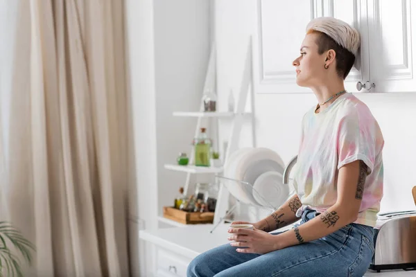 Joven mujer tatuada sentada en la encimera en la cocina y mirando hacia otro lado - foto de stock