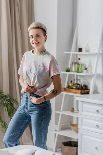 Mujer tatuada alegre mirando a la cámara mientras está de pie con la mano en el bolsillo en la cocina borrosa - foto de stock