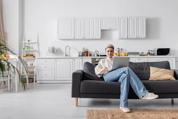 Повна довжина жінки в джинсах, сидячи на дивані з ноутбуком в сучасній кухні відкритого плану — стокове фото