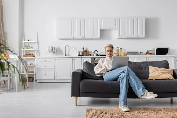 Tutta la lunghezza della donna in jeans seduta sul divano con computer portatile in cucina moderna a pianta aperta — Foto stock