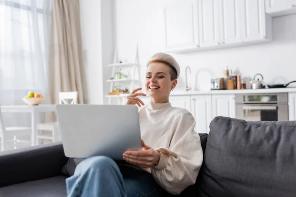 Excited woman with trendy hairstyle laughing near laptop on sofa at home — стоковое фото