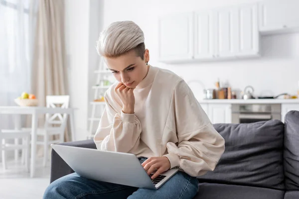 Pensive woman holding hand near chin while sitting on couch with laptop — Photo de stock