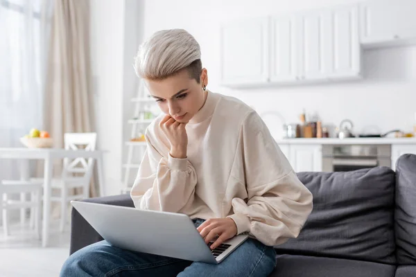 Pensive woman holding hand near chin while sitting on couch with laptop — стоковое фото