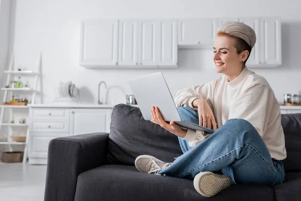 Donna felice guardando il computer portatile mentre seduto sul divano con le gambe incrociate — Foto stock