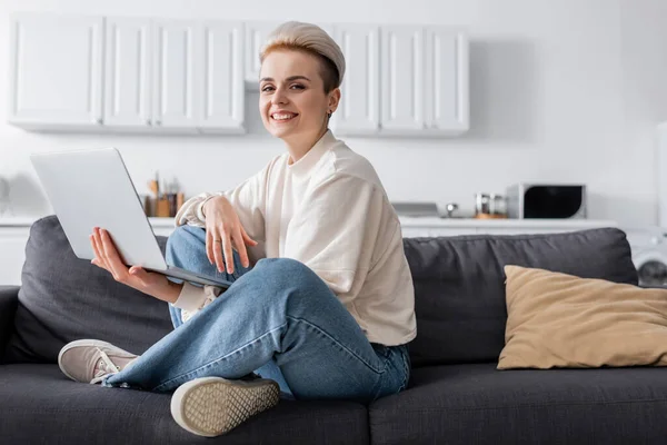 Frau mit Laptop sitzt mit überkreuzten Beinen auf Couch und lächelt in die Kamera — Stockfoto
