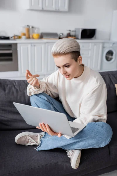 Модная женщина сидит на диване со скрещенными ногами во время видеозвонка на ноутбуке — стоковое фото