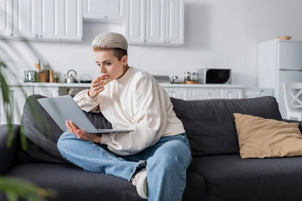 Thoughtful woman in white pullover sitting on sofa and looking at laptop — Photo de stock