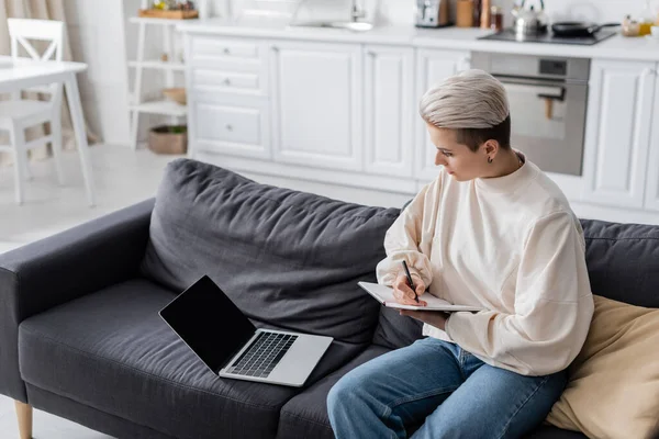 Femme avec une coiffure à la mode écrivant dans un ordinateur portable près du canapé à la maison — Photo de stock