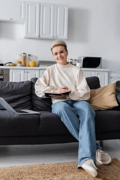 Полный рост женщины, сидящей на диване с ноутбуком и ноутбуком и улыбающейся перед камерой — стоковое фото