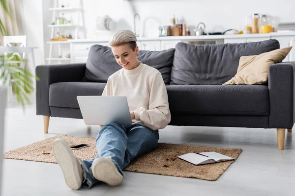 Full length of woman with laptop sitting on floor near couch, notebook and mobile phone — Stock Photo