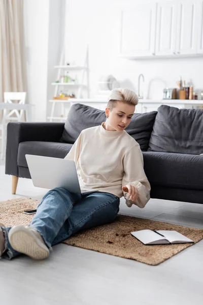 Trendy woman sitting on floor with laptop and looking at empty notebook — Foto stock