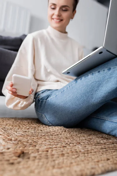 Mujer borrosa mirando el teléfono inteligente mientras está sentado en el suelo con el ordenador portátil - foto de stock