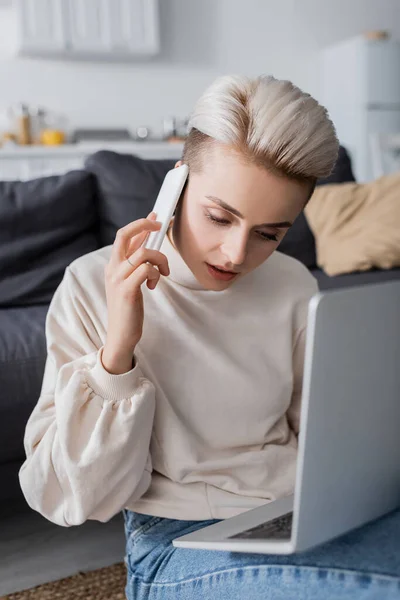 Donna in pullover bianco che chiama sul cellulare vicino a laptop sfocato — Foto stock