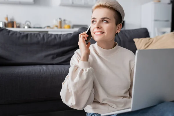 Cheerful woman looking away while talking on smartphone near laptop - foto de stock