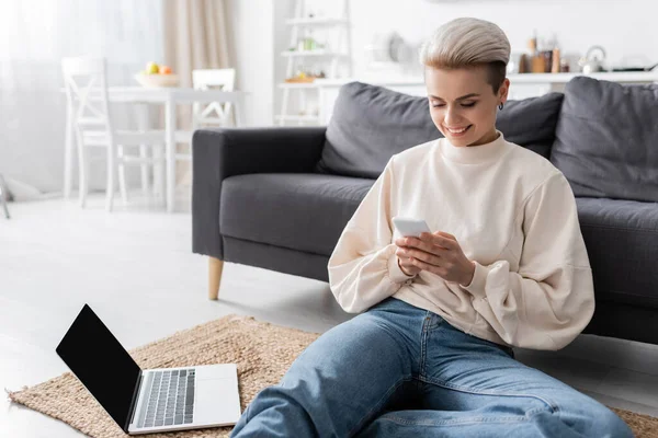 Щаслива жінка сидить на підлозі з мобільним телефоном біля ноутбука з порожнім екраном — стокове фото