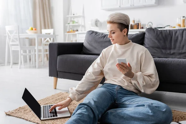 Positive woman with mobile phone sitting on floor and using laptop with blank screen — Foto stock