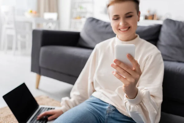 Cheerful woman sitting on floor near laptop and looking at mobile phone, blurred background — Photo de stock