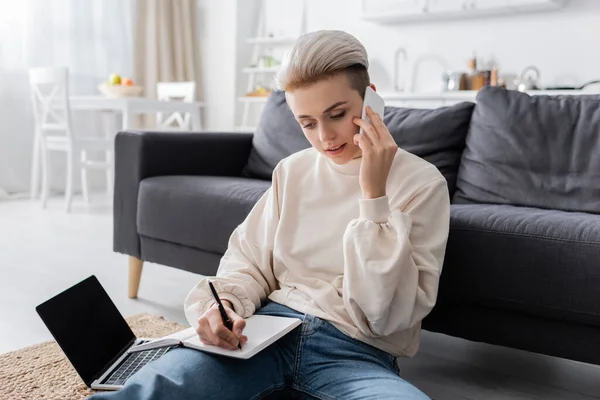 Young woman talking on smartphone and writing in notebook near laptop with blank screen — стоковое фото